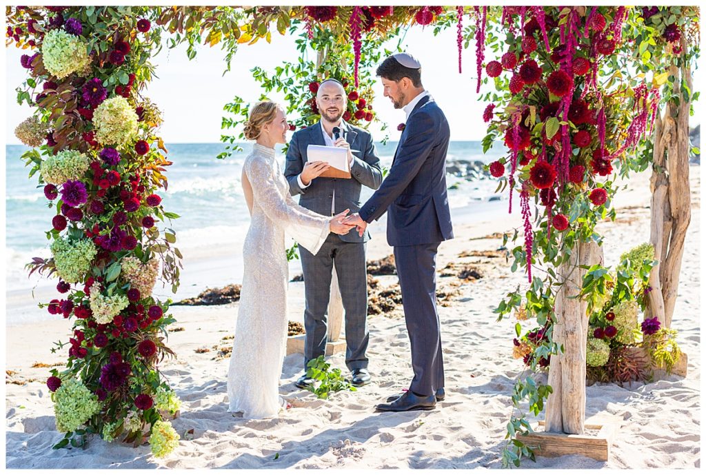 Bride and groom hold hands under floral chuppah on inn beach at their weekapaug wedding. 