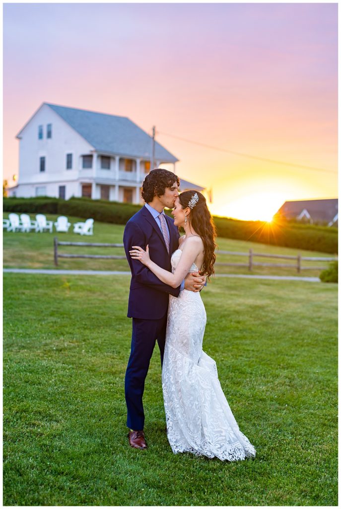 groom kissing bride on forehead during sunset with farmhouse in background