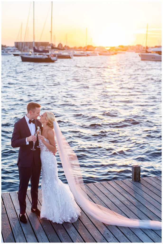 bride and groom cheers with champagne standing on dock with sun setting behind them