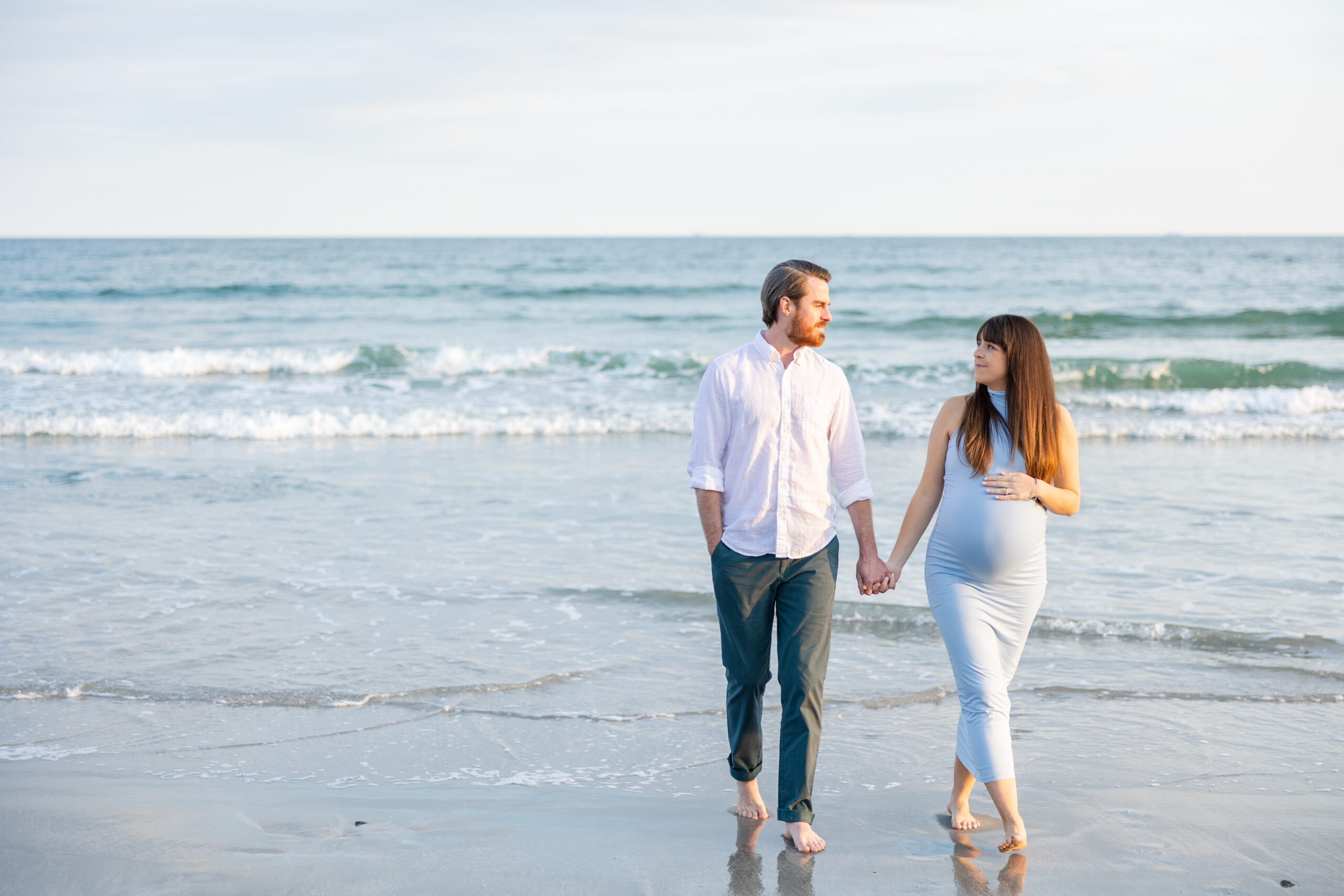 Pregnant wife and husband walk on sand during beach maternity session